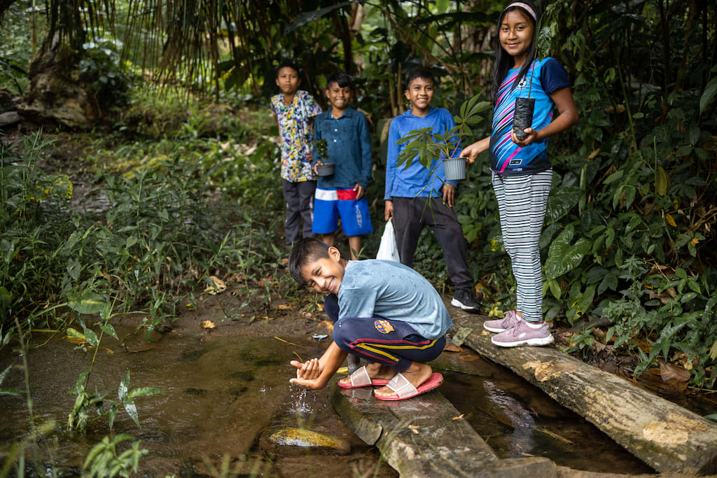 Martin is washing his hands in the water. Keysha is standing on a log behind him and holding a plant. His other friends are standing on the shore next to them.